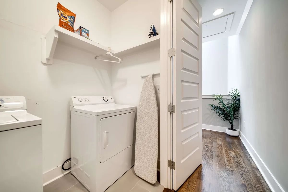 A dedicated laundry area with a washer, dryer, ironing board, and shelving. The setup is organized and functional, conveying a sense of efficiency and daily ease for residents or guests managing laundry needs during their stay.
